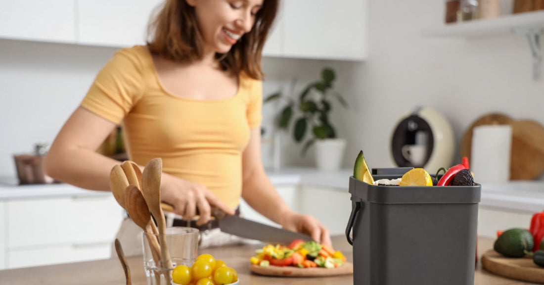 A woman cutting vegetables on a wooden counter beside a gray compost bin, avocados, onions, tomatoes, and utensils.