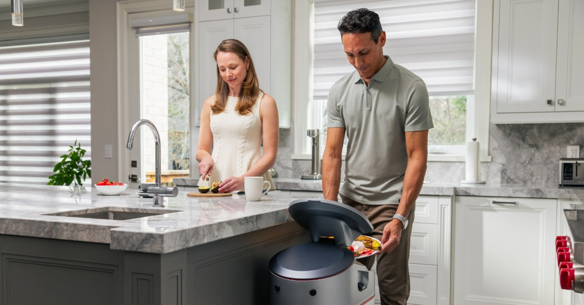 A man places food scraps into a kitchen waste machine while a woman prepares produce in a bright kitchen.