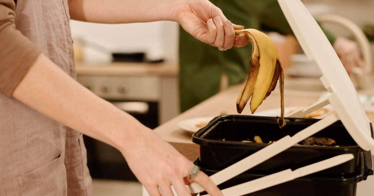 Inside a kitchen, a person holds a banana peel above an open black compost bin with a white lid near a countertop.