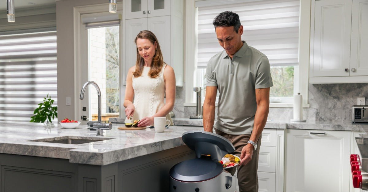 A woman slicing avocado while a man discards food scraps into a kitchen bin beside a marble countertop.