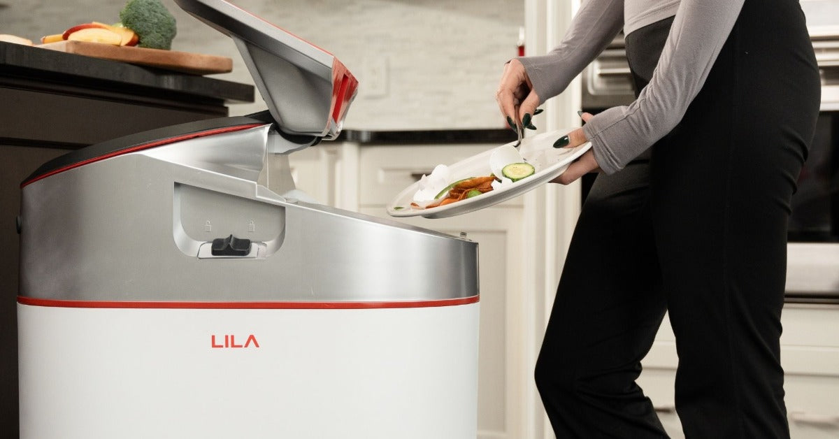 A person scraping food scraps from a plate into an open LILA composter appliance beside kitchen cabinets.