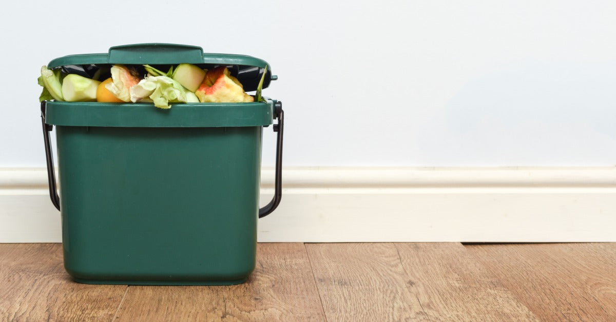 A green compost bin filled with fruit and vegetable scraps sitting on a wood floor against a pale wall.